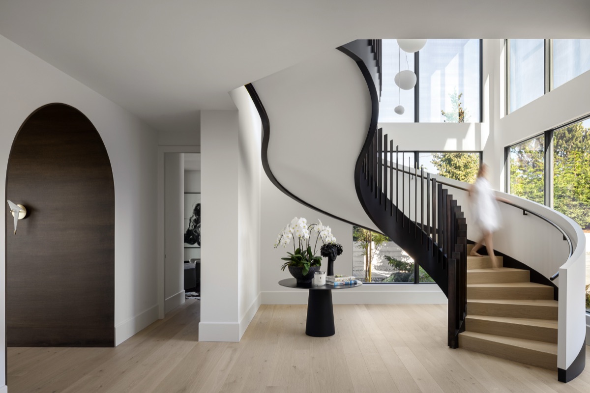 A curved black stair rising through a double-height entry beside a wall of glass