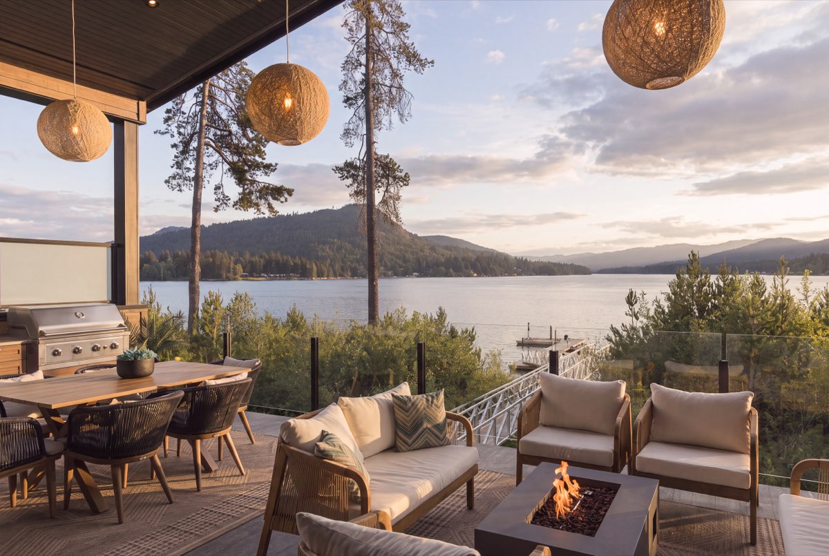 A cedar deck with woven pendant lanterns and low seating above a still inlet at dusk.