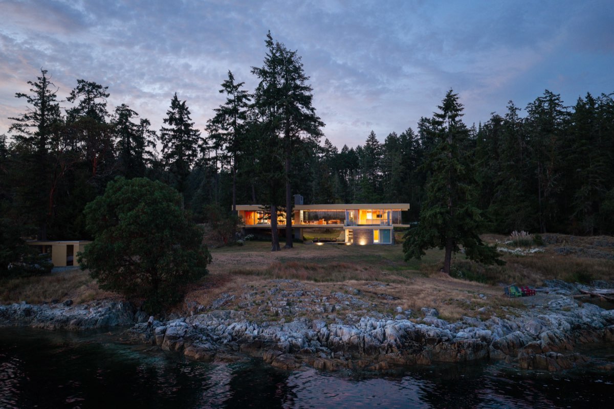 A low house lit at dusk across an open meadow, the treeline behind.